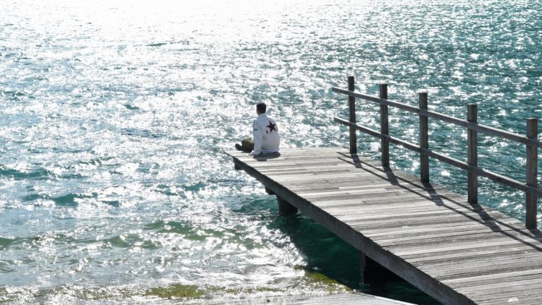 A person sits on a wooden pier, gazing at a shimmering lake with mountains and a sailboat in the background.